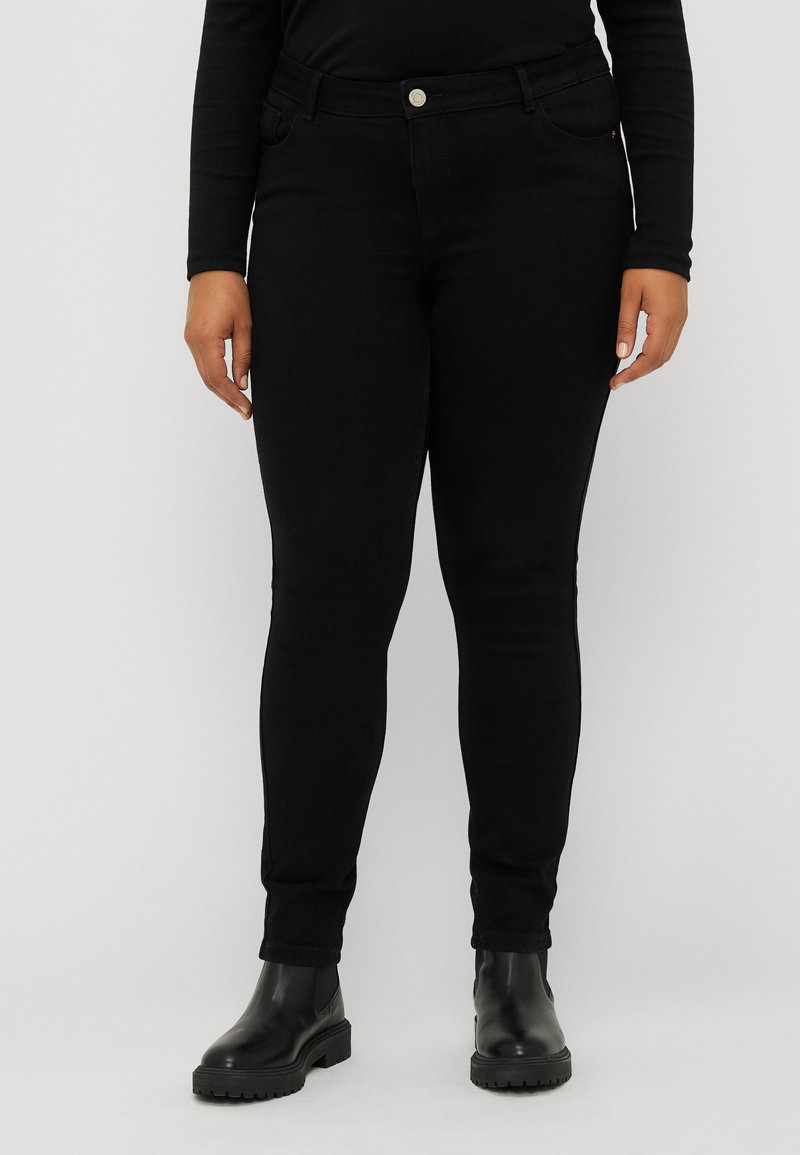 Jean skinny noir avec une texture lisse, doté de poches avant, d'une fermeture à bouton, et d'un design ajusté. Porté avec des bottes noires épaisses.