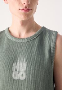 Bezrukávový zelený rebrovaný top s okrúhlym výstrihom a jemnou bielou grafikou na hrudi. Materiál vyzerá mäkko s textúrovaným povrchom.