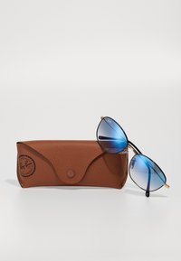 Ray-Ban'i prillid siniste gradiendi läätsedega, kuldse metallraamiga ja mustade tempelkätega, puhkavad pruuni tekstuuriga nahast kotikese kõrval.