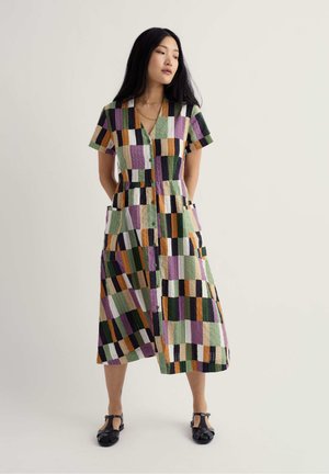 Vrouw staat met handen achter de rug, draagt een jurk met korte mouwen en knoopsluiting, met patchwork ruitmotief, en zwarte sandalen met gesloten tenen.