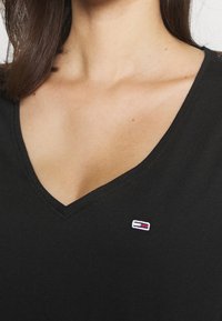 Svart V-ringad t-shirt med en figurnära design. Har en liten broderad logotyp i röd, vit och blå på den övre vänstra sidan. Mjuk tygtextur.