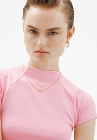 Top ajustado rosa con un cuello alto, mangas cortas y textura suave. Acentuado con collares en capas: uno de oro con símbolo de infinito y otro de plata con un colgante.