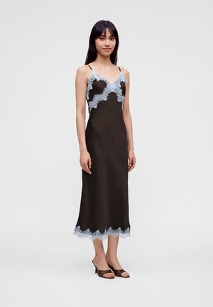 Frau trägt ein schwarzes Spaghettiträgerkleid mit blauen Spitzendetails am Ausschnitt und Saum, steht vor einem schlichten hellen Hintergrund.