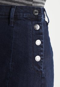 Tmavě modrá džínová sukně s měděnými krycími knoflíky na boku, s hladkou texturou a klasickým těsným střihem.