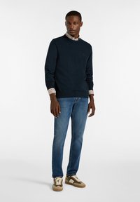 Dunkelblauer Pullover mit rundem Hals, strukturiertem Stoff, kombiniert mit einem Hemd mit Kragen und gestreiften Akzenten. Trägt dazu blaue Jeans und beigefarbene Sneaker.
