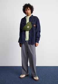 Giacca di jeans, camicia verde chiaro, pantaloni grigi e sneaker beige. Marsupio verde con logo, indossato trasversalmente sul petto. Capelli ricci, sfondo neutro.