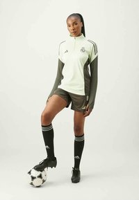 Zöld és szürke Adidas edzőfelső negyed cipzárral, rövidnadrág és fekete zoknik. A modell egy focilabdán áll, csíkos részletekkel.