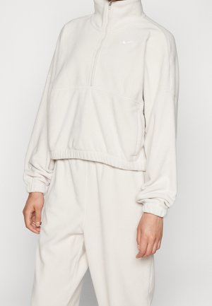 Model draagt een witte fleece pullover met een ritssluiting bij de kraag en bijpassende fleecebroek, handen ontspannen langs het lichaam.