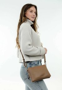 Donna in un maglione grigio chiaro a collo alto e jeans blu che porta una borsa a tracolla in pelle marrone su sfondo bianco.