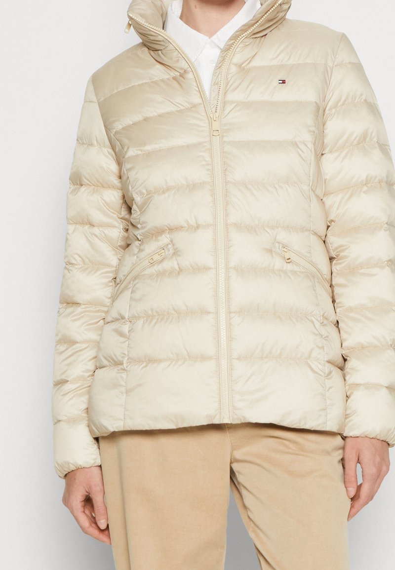 Persona vistiendo una chaqueta acolchada beige con cremallera frontal y bolsillos con cremallera, combinada con pantalones marrón claro.