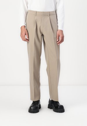 Bežid kohandatud püksid sirge lõike ja esikortsudega. Valmistatud kergest kangast, koos mustade massiivsete taldadega kingadega.