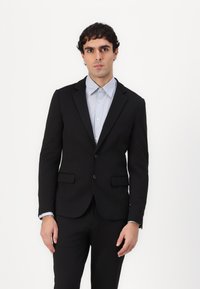 Schwarze taillierte Sakkjacke mit zwei Knöpfen, Reverskragen und vorderen Taschen, kombiniert mit einem hellblauen Hemd mit Knopfleiste.