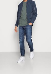 Giubbotto bomber della marina con zip, camicia verde sottostante, jeans blu e sneaker bianche. Il giubbotto presenta una tasca laterale e polsini a coste.