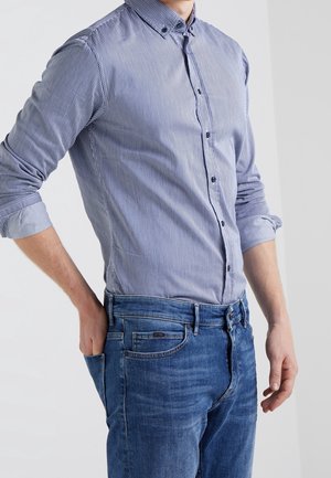 Mand iført en blå og hvidstribet skjorte med knapper, med ærmerne rullet op og blå jeans, hånd i baglommen, stående mod en ensfarvet baggrund.