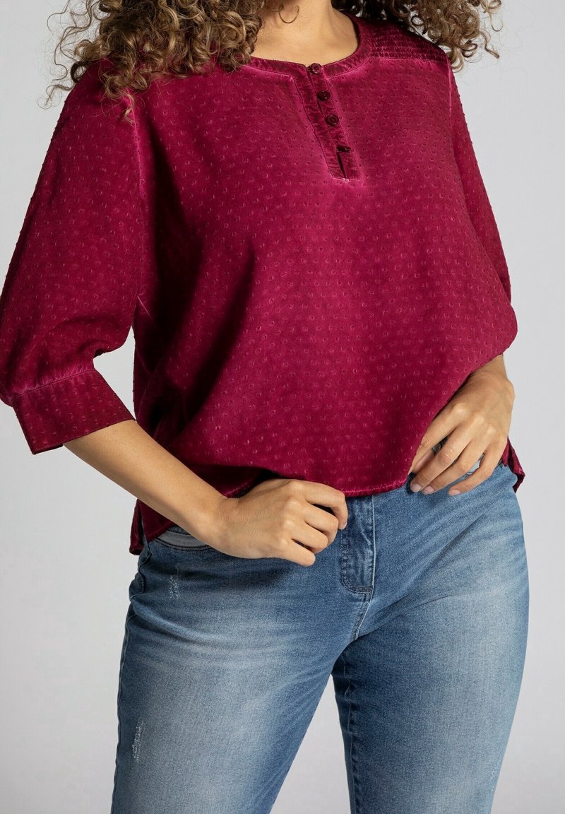 Blouse bordeaux en tissu texturé, col rond, trois boutons et manches trois-quarts. Associée à un jean en denim bleu clair.