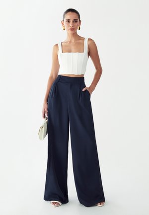 Hvid cropped top med teksturerede paneler og brede stropper, kombineret med marineblå vidde bukser med plisseringer, holdt oppe med et sort bælte.