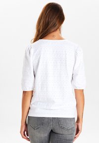 White short-sleeve top with a textured pattern and subtle dot accents. Features a relaxed fit and gathered sleeves. Paired with gray jeans.