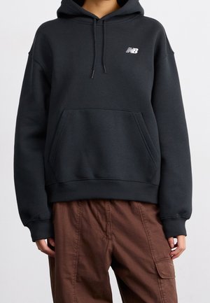 Osoba nosząca czarną bluzę z kapturem New Balance z przednią kieszenią i sznurkami, w połączeniu z brązowymi spodniami typu cargo.