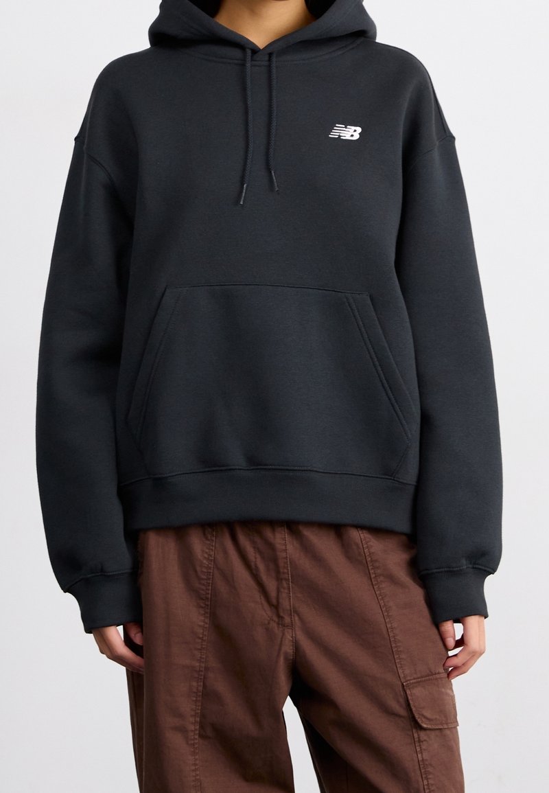 Personne portant un sweat à capuche noir New Balance avec poche avant et cordons, associé à un pantalon cargo marron.