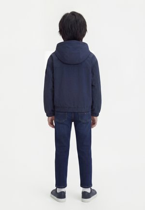 Kind steht mit dem Rücken zur Kamera, trägt eine marineblaue Kapuzenjacke, dunkelblaue Jeans, weiße Socken und marineblaue Turnschuhe vor einfarbigem Hintergrund.