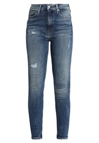 Vaqueros de mezclilla azul de corte Slim-fit, con áreas desgastadas, parches rasgados y cierre de botón. Presenta cinco bolsillos y una textura ligeramente usada.
