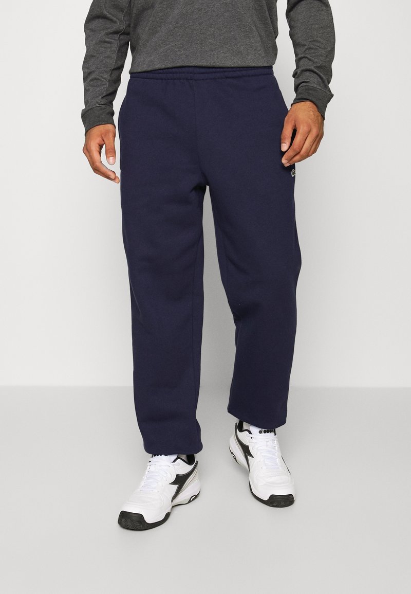 Navy fleece sweatpants with an elastic waistband, straight cut, and a small logo detail on the left thigh. Worn with white athletic shoes.