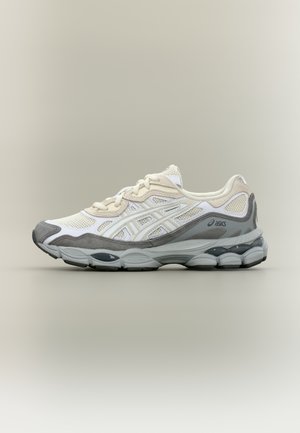 Zapatilla deportiva blanca y gris con parte superior de malla, cuello acolchado y una suela gruesa y acolchada que presenta un detalle y logo en gris.