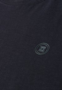 Tissu noir avec un petit logo circulaire comportant une lettre G stylisée à l'intérieur, placé près de la partie supérieure de la poitrine.
