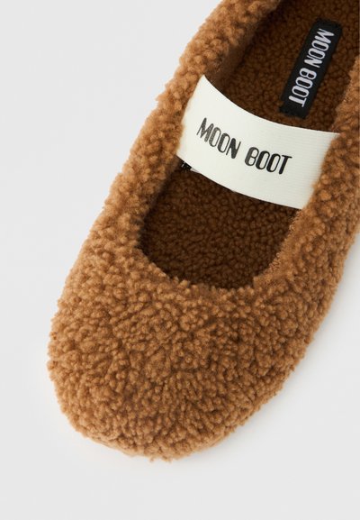 Chausson marron duveteux avec une lanière blanche sur le dessus portant l'inscription "MOON BOOT" sur un fond blanc.