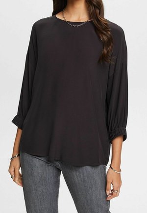 Nainen, joka pukeutunut löysään mustaan pitkähihainen toppiin, jossa on kerätyt hihansuut, hopeinen ketjukaulakoru, sormuksia ja harmaat farkut yksinkertaisen taustan edessä.