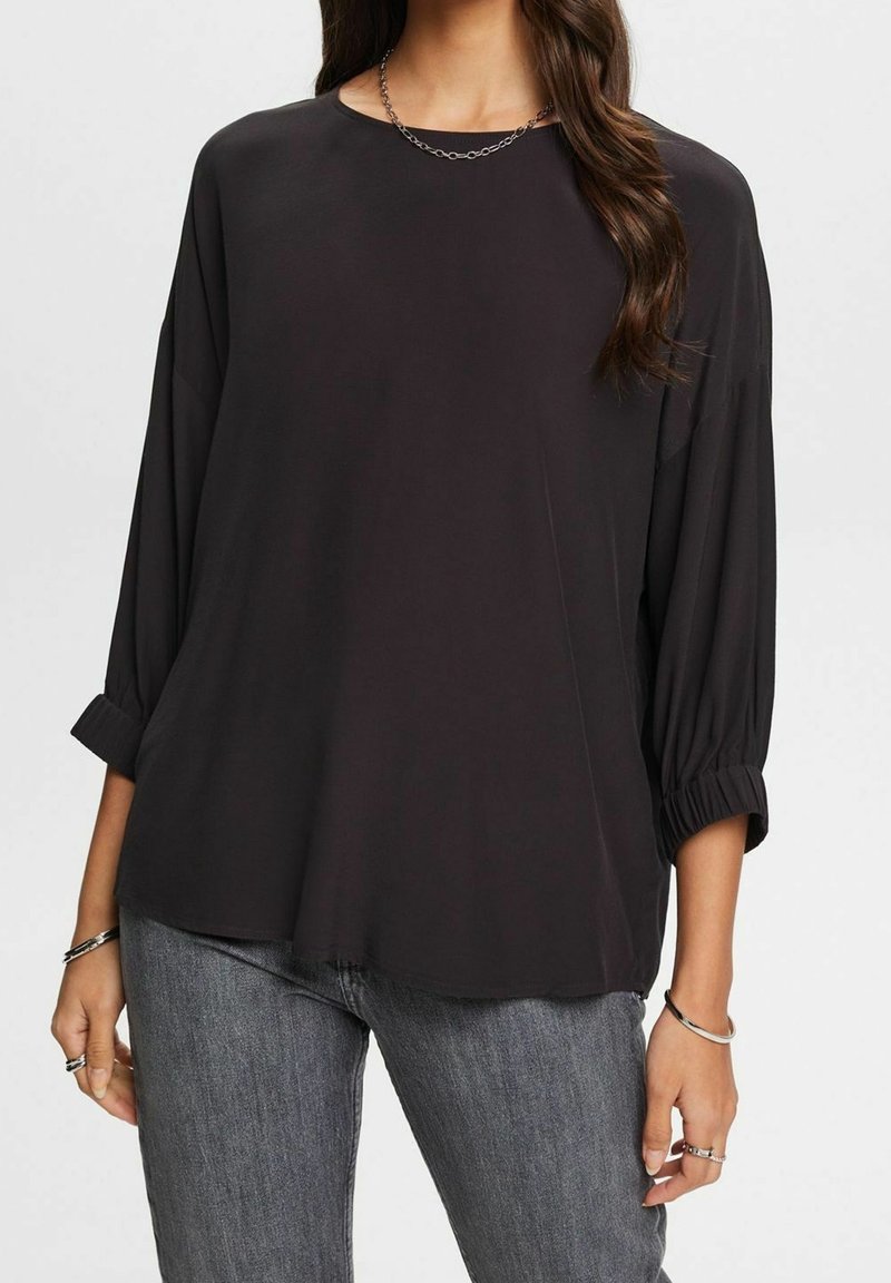 Femme portant un haut ample noir à manches longues avec poignets froncés, un collier en chaîne argentée, des bagues et un jean gris, sur un fond uni.