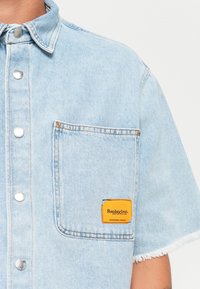 Lyseblå denim skjorte med kortærmer, sølvfarvede trykknapper, en brystlomme og et orange mærke, der viser "Replay Inc." ved kanten af lommen.