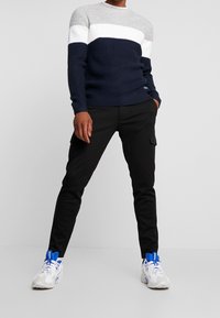 Man wearing color-block gray, white, and navy knit sweater, black cargo pants, and white sneakers with blue accents, standing with hands partly in pockets.