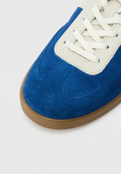 Modrá semišová teniska s béžovým koženým jazykom, vybavená bielymi šnúrkami a textúrovanou hnedou gumovou podrážkou. Mäkká textúra a športový dizajn.