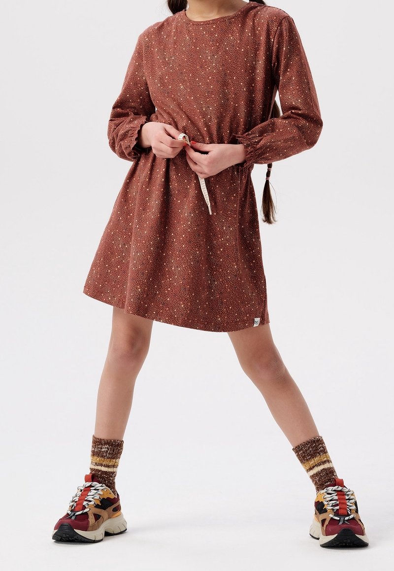Enfant portant une robe à manches longues couleur rouille avec de petits pois floraux, tenant un mètre ruban, chaussant des baskets colorées et des chaussettes à motifs.