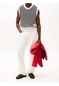 Czarnobiały sweter w paski bez rękawów, białe dżinsy, trzyma czerwoną pikowaną kurtkę. Outfit dopełniają czarne mokasyny.