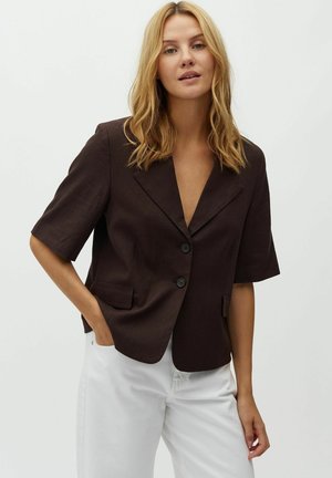 Blond žena v tmavohnedom krátkom saku s dvomi gombíkmi a bielych nohaviciach pózujúca na jednoduchom svetlom pozadí.