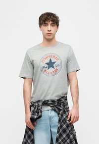 Νεαρός άνδρας με κυματιστά μαλλιά, φορώντας γκρι μπλουζάκι Converse All Star, ανοιχτό μπλε τζιν και μαύρο καρό πουκάμισο δεμένο γύρω από τη μέση του.