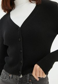 Czarny prążkowany kardigan z dekoltem w kształcie V, z przodem zapinanym na guziki i długimi rękawami. Nosi się go na białej prążkowanej bluzce o fakturowanym wykończeniu.
