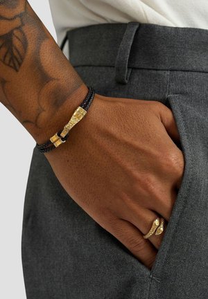 Sort flettet armbånd med en struktureret guldlås, båret på et håndled, kombineret med grå bukser. Subtil hudtekstur synlig.