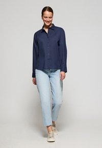 Marineblaues Hemd mit langen Ärmeln, aus leichtem Stoff und in lockerer Passform, kombiniert mit hellblauen geraden Jeans und beigen Sneakers.