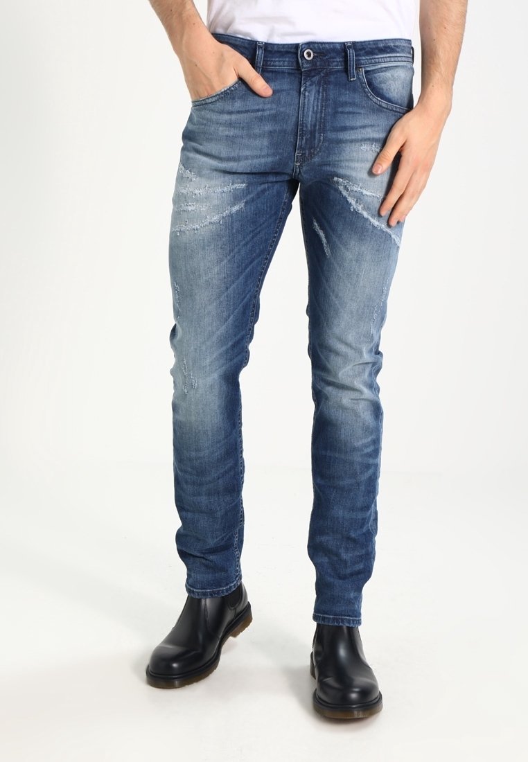Homme portant un jean slim bleu délavé avec des détails usés, des bottines en cuir noir et une chemise blanche, debout les mains près des poches.