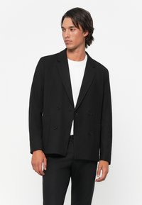 Blazer negro de corte cruzado hecho de una tela suave, con dos bolsillos frontales, botones grandes y un diseño clásico de solapa.