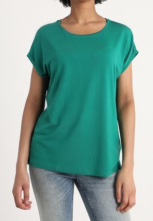 Kobieta w luźnej, morskozielonej bluzce z krótkim rękawem wywiniętym na mankiet oraz w jasnoniebieskich dżinsach na jednolitym tle.