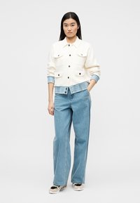 Γυναίκα που στέκεται φορώντας λευκό σακάκι με κουμπιά πάνω από μπλε πουκάμισο με σχέδια, φαρδιά μπλε τζιν και μπεζ αθλητικά παπούτσια με μαύρες λεπτομέρειες.