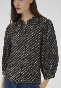 Fekete és szürke átlós csíkozású blúzt viselő nő, puffos hosszú ujjakkal és gombos mandzsettával, kék farmernadrággal párosítva.