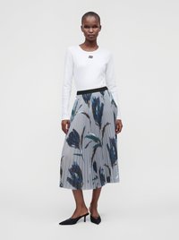 Femme portant un haut blanc à manches longues et une jupe midi plissée grise avec de grands imprimés floraux bleus, associée à des mules noires à talons.