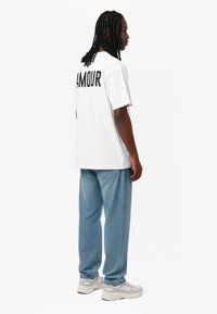 Bijela oversized majica s natpisom "AMOUR" u crnoj boji na leđima, u kombinaciji s svijetloplavim trapericama i bijelim sportskim tenisicama.