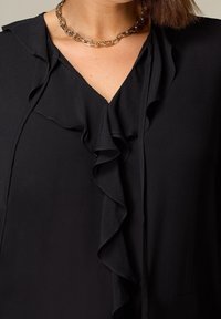 Svart blus med V-ringning, vertikala volanger framtill och ett mjukt, strukturerat tyg. Guldhalsbandet framhäver dekoltén.