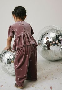 Lila Samt-Outfit mit kurzen Ärmeln, Peplum-Taille und Schleifenmustern, kombiniert mit passenden Hosen, neben zwei großen Discokugeln.